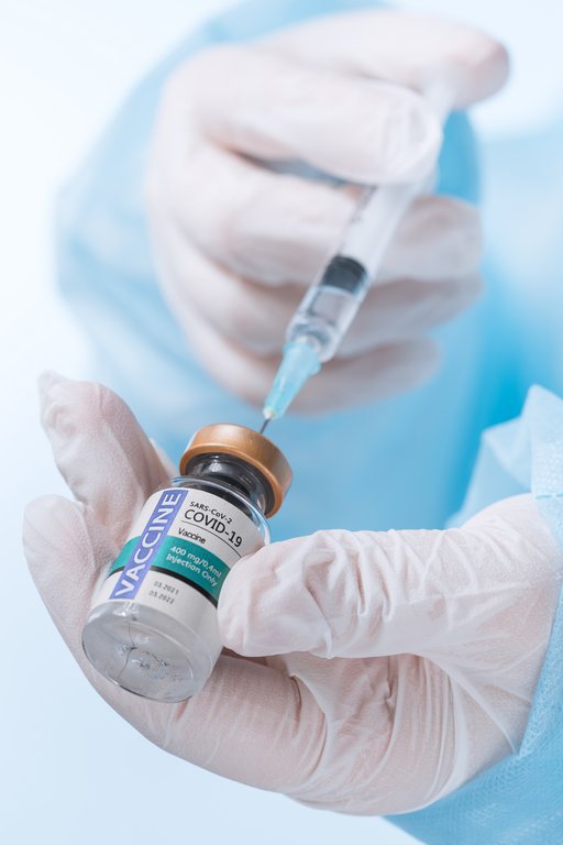 Ett par medicinskt handskklädda händer håller ett covid-19-vaccin med en kanyl i. Vätska sugs upp från den lilla flaskan.