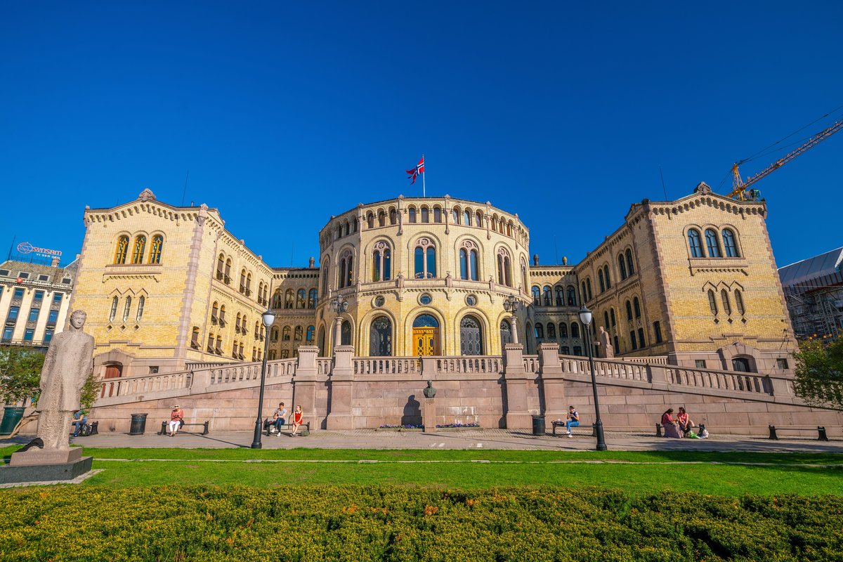 A symmetrical view of the Norweigan parliament in Oslo. The building has one round building flanked by two square buildings. The background is a pure blue sky, and green grass is in front of the parliament.