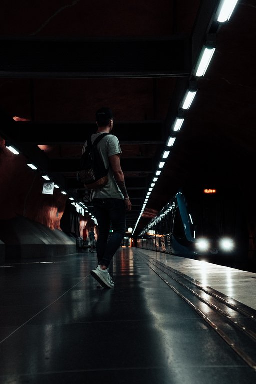 A boy is standing in a subway in Stockholm. The photo is very dark, with bright lines of light in the ceiling. A train is at the platform, and the boy seems to be heading for the train.