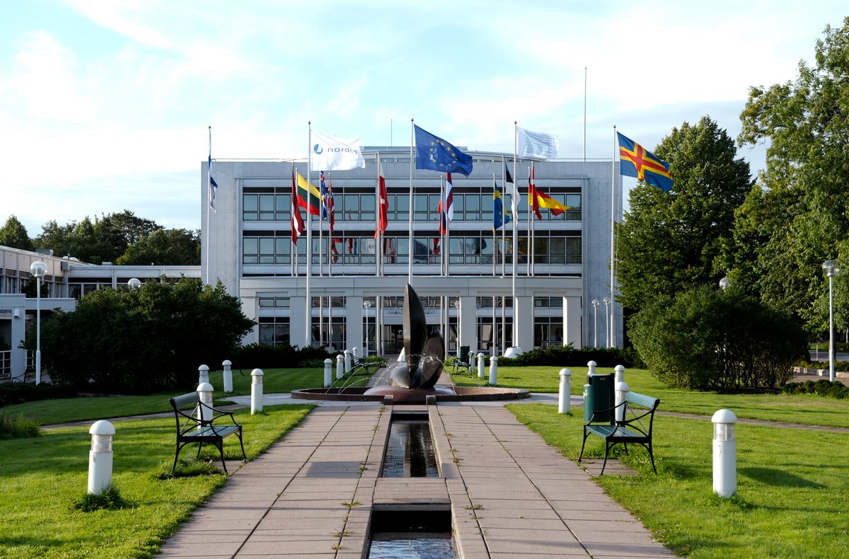 The entrance of the Parliament of Åland. There is a path to the entrance with green grass on each side. There are all the Nordic flags. The building itself is rectangular, white and with glass windows.