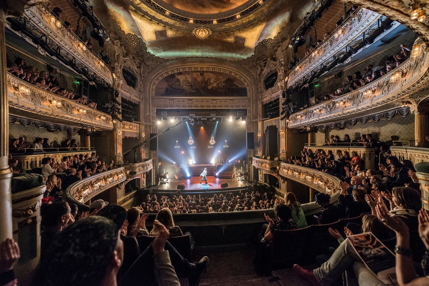 Inside of a theatre, large round shaped seatings with a large crowd looking at the scene. The hall is filled with lights and colours.