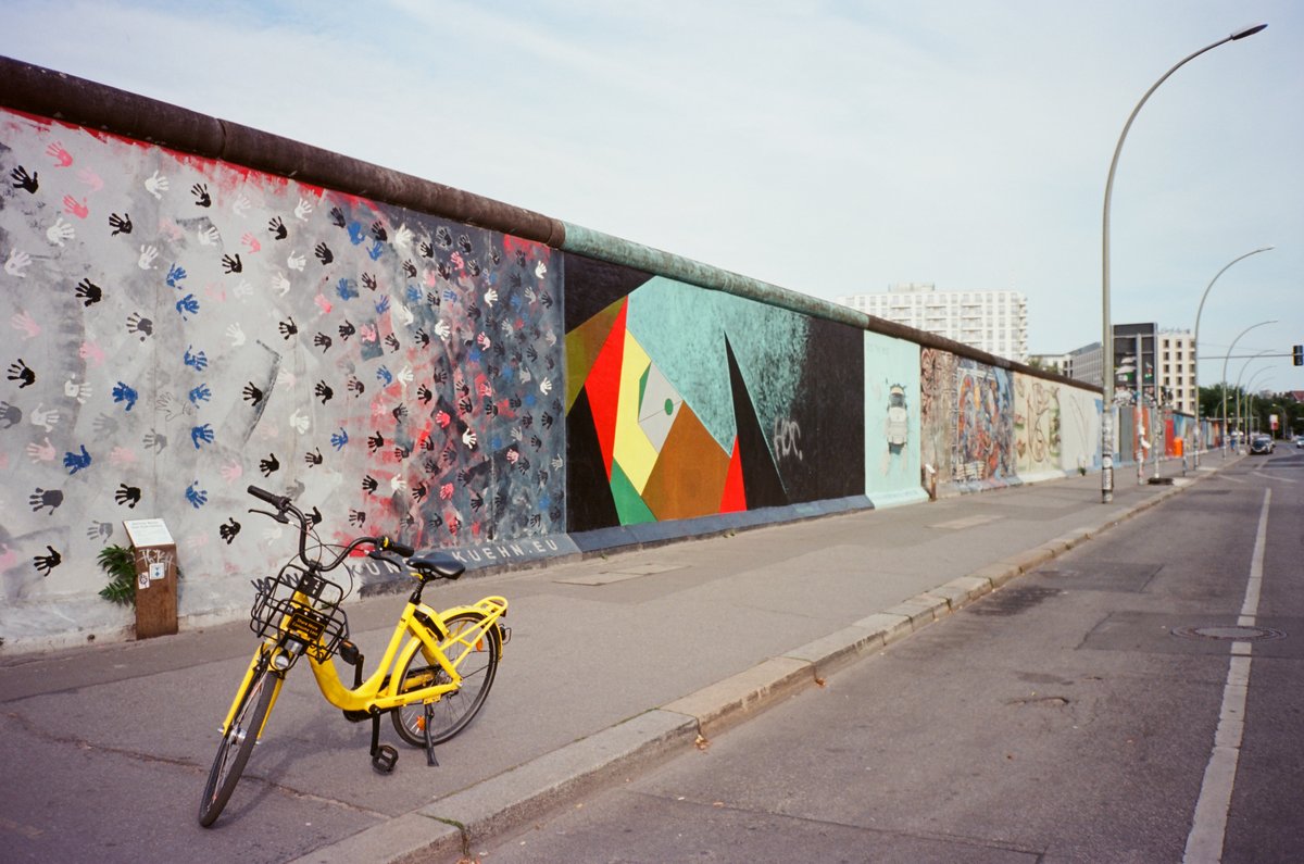 På billedet ses rester af Berlinmuren, dekoreret med farverig graffiti af håndaftryk og moderne, abstrakt kunst. En lysegul bil er parkeret foran muren.