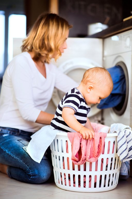 A woman is sitting on the floor in front of a washing machine with a baby sitting in a clothes basket next to her. She is removing clothes from the machine.