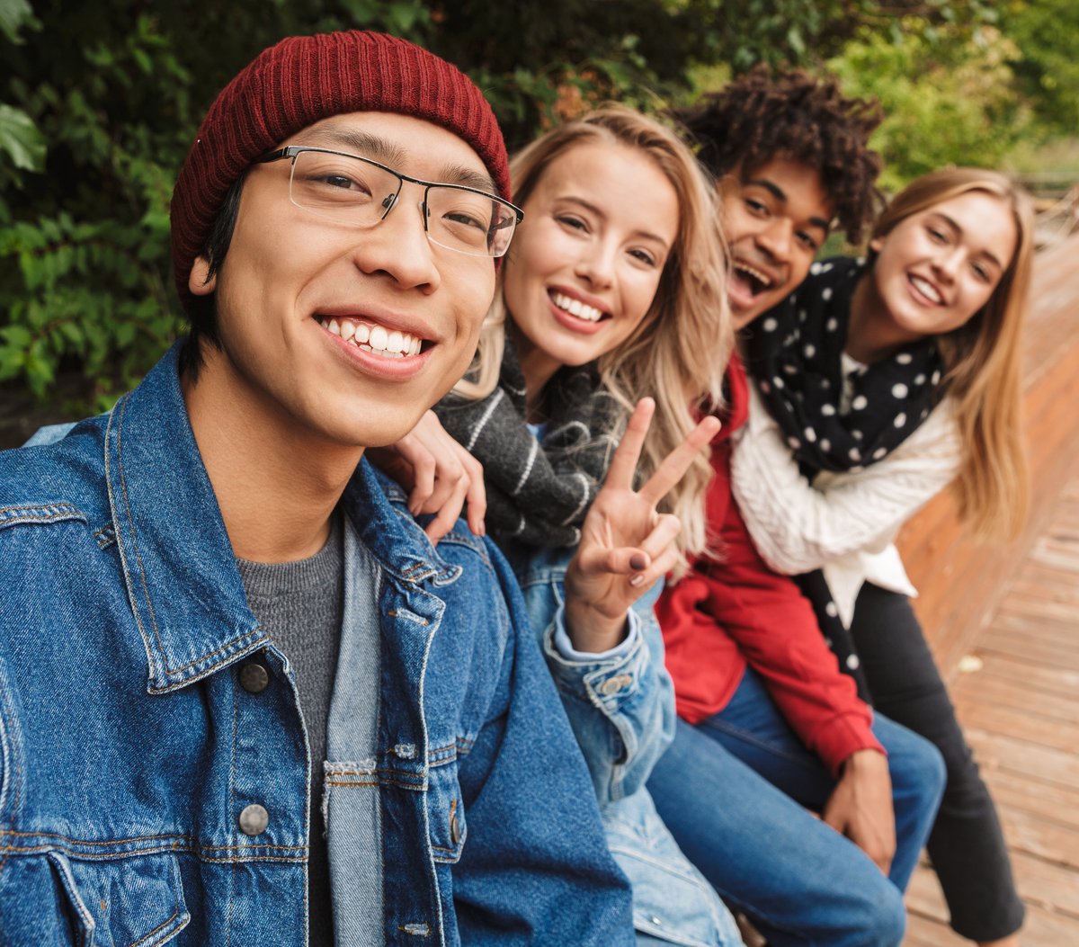 A picture of four young people sitting beside each other on red bricks besides a green bush. They are all smiling and looking at the camera. One of the girls is holding up her hand, making the peace sign with her fingers.