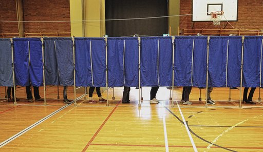 På billedet ses danske stemmebokse i en gymnastiksal med blå gardiner trukket for. Kun benene på dé personer, der er inde og stemme, er synlige under gardinet.