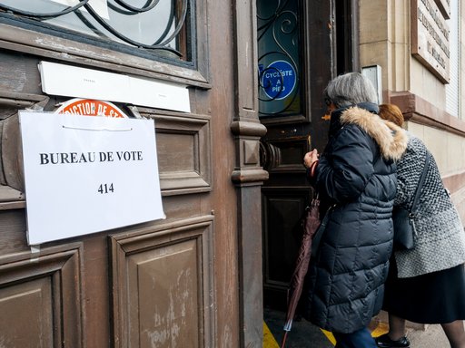 Two elderly women are entering a polling place. They are entering through a thick wooden for, on which a sign is hanging saying "Polling place" in French.