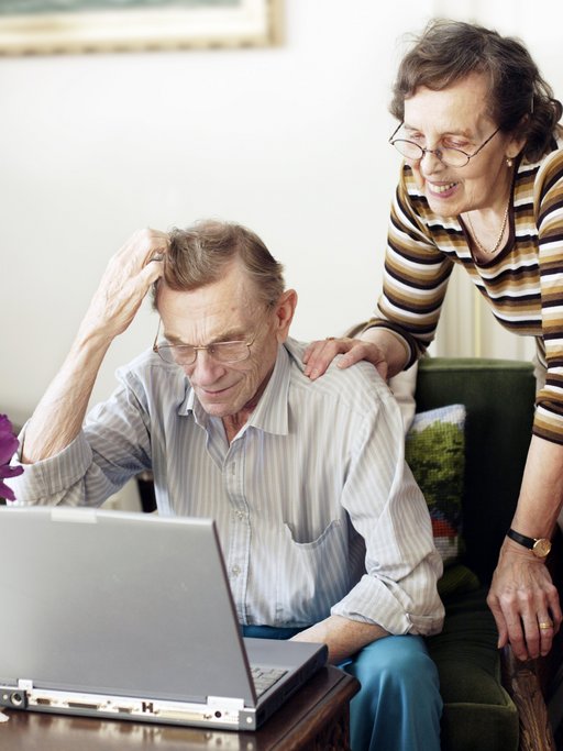 A picture of an old man sitting down in front of a computer. His hand is scratching his head, indicating that he is confused or upset. Behind him, an elderly woman is smiling and has a hand on his shoulder.