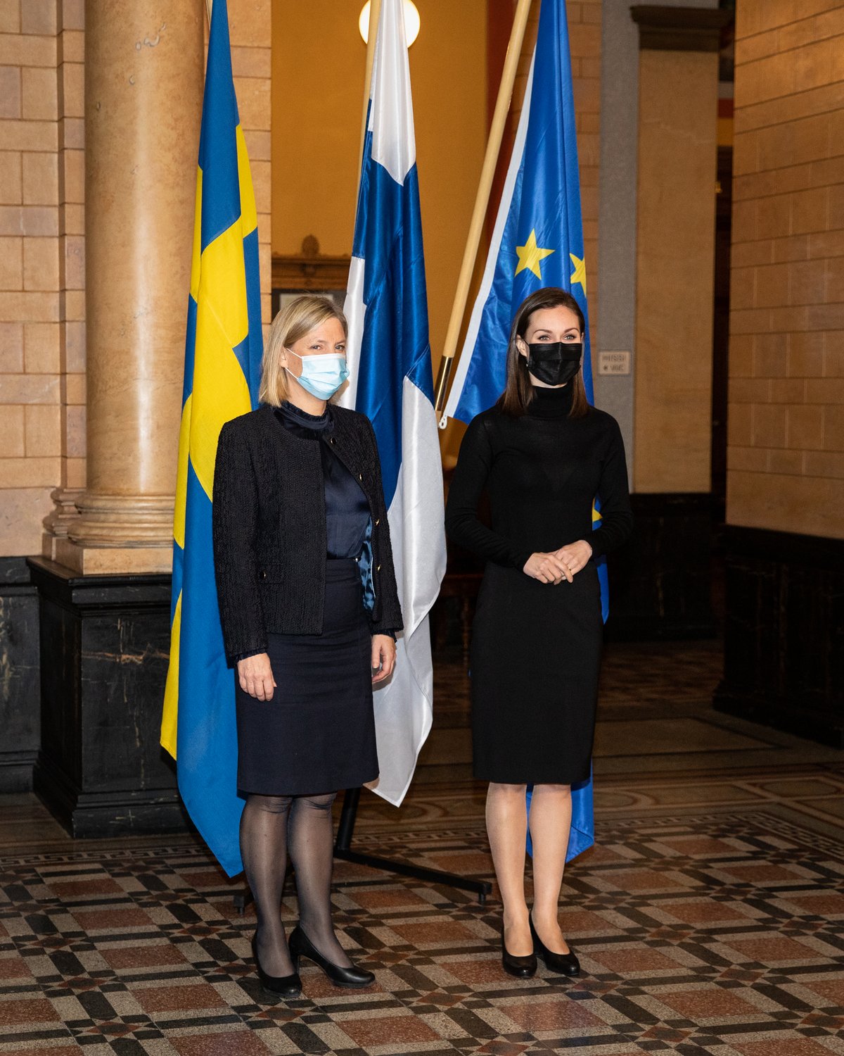 The Finnish Prime Minister is standing next to the Swedish Prime minister with their respective flags behind them as well as a flag for the EU. They are both wearing face masks.