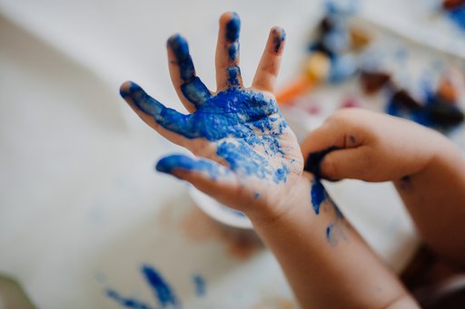 Pictured is a child's hand held forth, covered in blue paint. In the blurred background, other painting supplies can be seen.