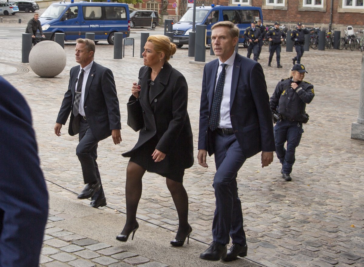 Inger Støjberg and Kristian Thulesen Dahl walking besides one another on a cobblestone street. Støjberg is looking to the side away from the camera and Thulesen Dahl is looking at the camera. In the background are two police cars and several police officers, most likely escorting the two politicans somewhere.