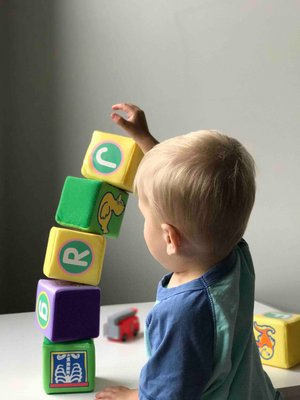 A picture of a young, blonde boy seen from behind. He is playing with colourful cubes with different letters and pictures on them, having stacked them into a small tower.