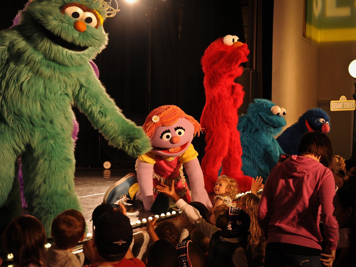 Kuvassa on Katie-niminen Sesame Street-hahmo, joka kurkottaa esityksen aikana kohti yleisössä olevia lapsia. Lapset näyttävät onnellisilta ja kurkottelevat ylöspäin kohti kookasta nukkea.