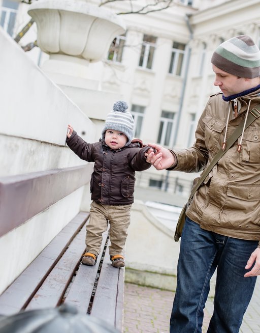 A young child is playing around on a bench, holding the hand of a man, presumably the baby's dad.