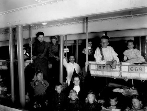 Black and white photo of immigrants sitting next to each other on bunkbeds, on a ship. 