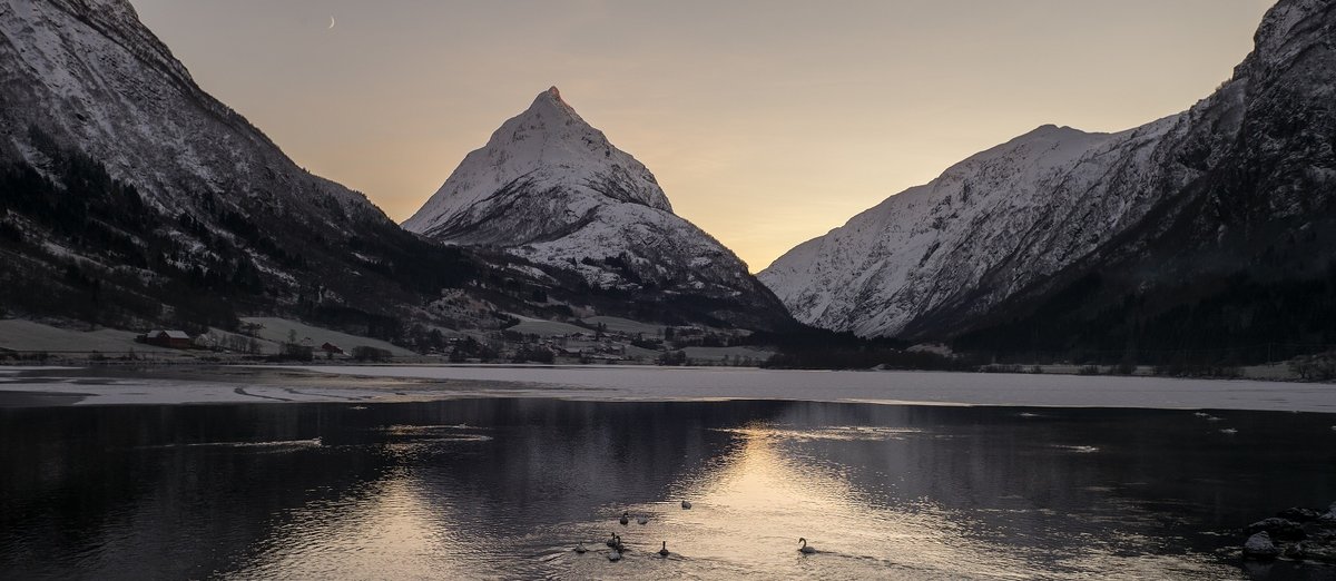 A picture of a rocky mountain with a snowy peak against a clear sunset sky. In front of the mountain is a lake half frozen and with birds swimming in it.