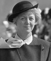Portrait of Vigdís Finnbogadóttir. Here, she is a young woman with blond short hair, standing upright wearing a black long trenchcoat and a formal hat.