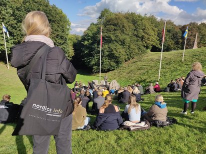 Nicola Witcombe standing with her back to the camera. She is looking at a hilly landscape, Skamlingsbanken in Kolding, Denmark. Several young people are sitting on the grass, listening to a speaker. Flags from the Nordic countries are scattered across the hills. Over her shoulder, Witcombe is wearing a black tote bag with the nordics.info logo printed across it.