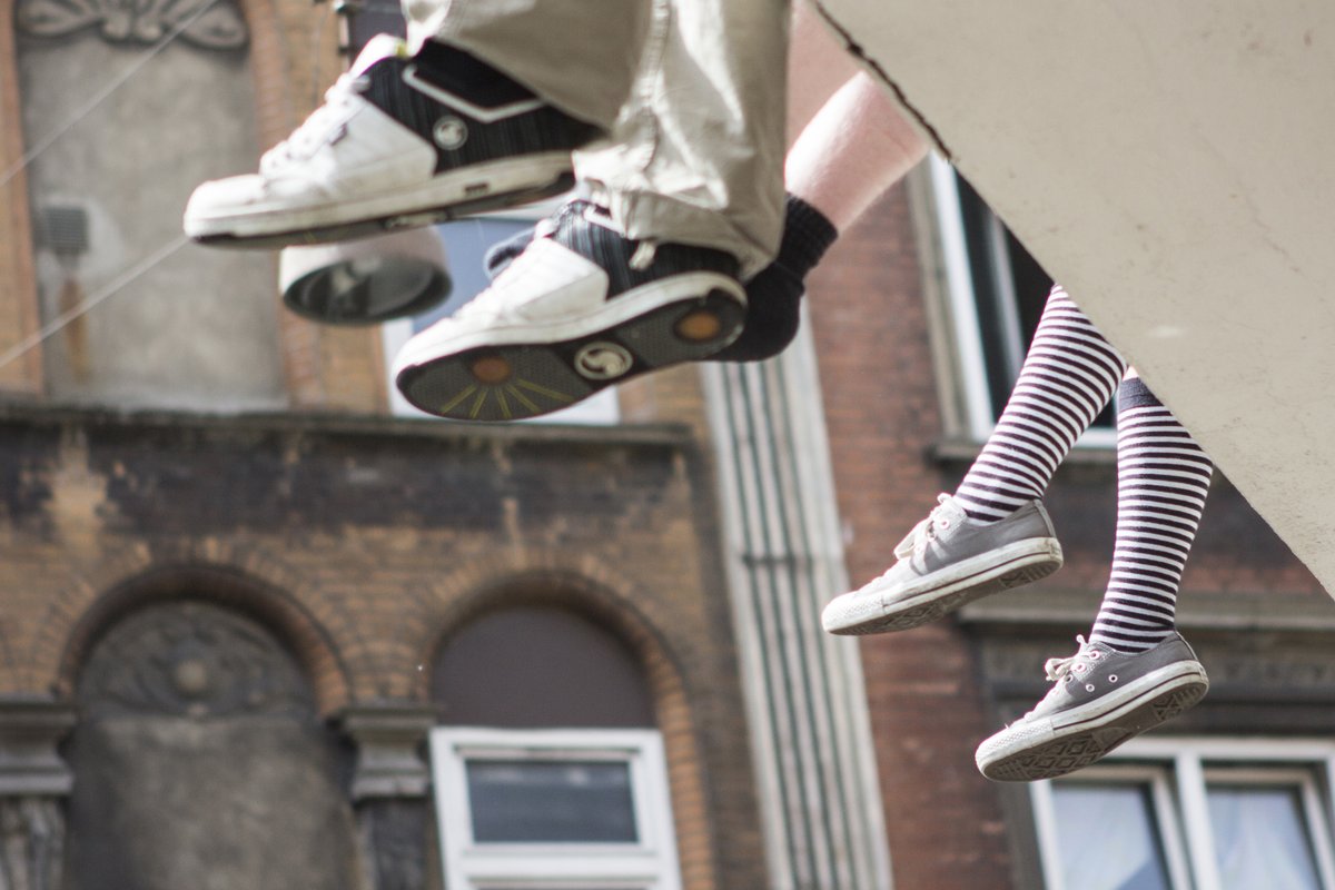 A picture of three pair of legs dangling below a concrete edge. From left to right, the legs are wearing jeans and sneakers, running shoes, and long knee-high striped socks with sneakers.