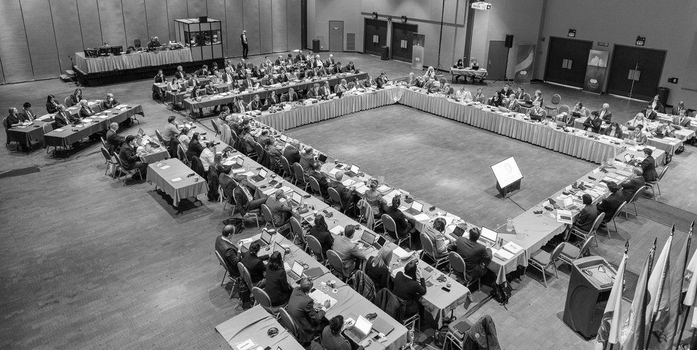 black and white photo of a meeting room. The tables are lined up in the shape of a square and there are people sitting and chatting
