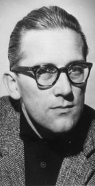 Black and white photo of a man with square glasses and slicked back hair. He is wearing a black shirt and a jacket