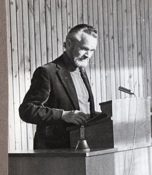 Black and white picture of a younger Niels stading at a podium, doing a speech. He seems to have grey long hair to his shoulders and a grey beard.