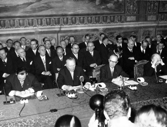 Black and white photo with a group of men in similar suits from 1950s sitting down with old-fashioned microphones and journalists at the front
