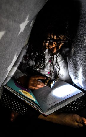 A child sitting under a blanket with a flashlight and is reading a book