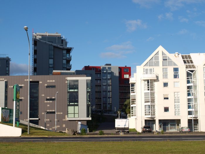 Horisontal picture of four quite different looking buildings. Building 1 is wide, beige brown. Building 2 is tall and thin, dark grey. Building 3 is square and light grey with red blocks. The last building is white and triangular.