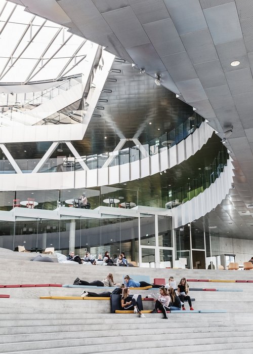 Inside the Tórshavn education centre. In the front, there is a big staircase where young people are sitting, chatting and lying down, relaxing. Behind the staircase, you can see that the building has several stories and at the top, the sunlight clearly sheds through. Inside of the building is with metal plates.