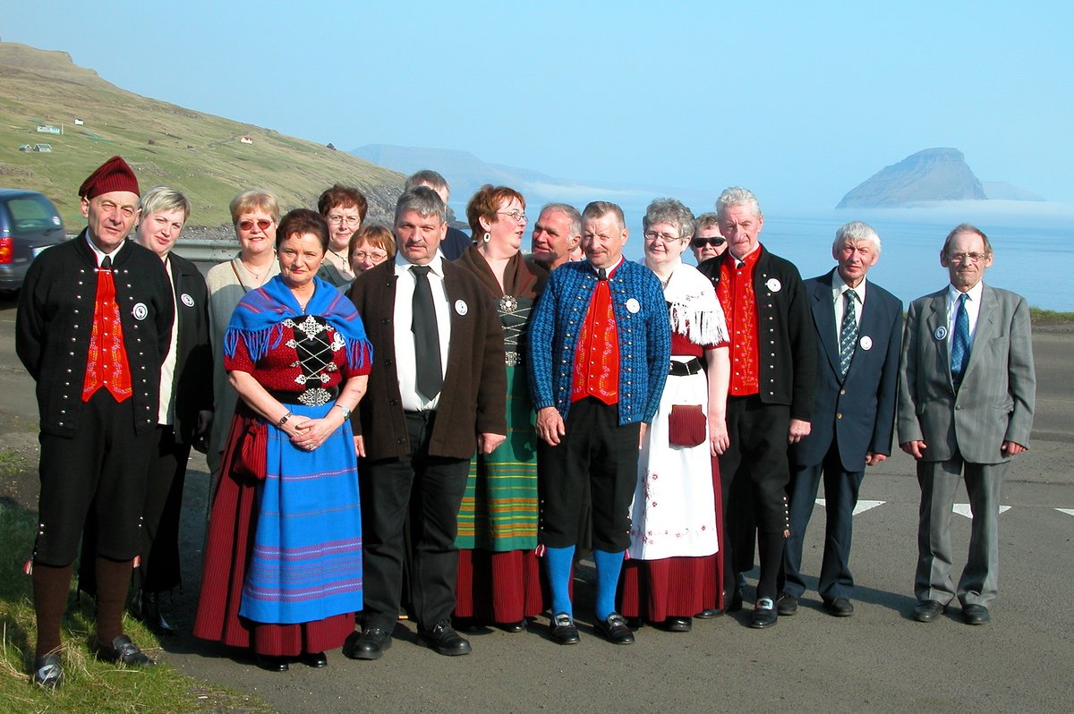 A photo of a group of people wearing traditional Faroese national clothing. In the background are mountains and the sea. The people are all looking at the camera and smiling.