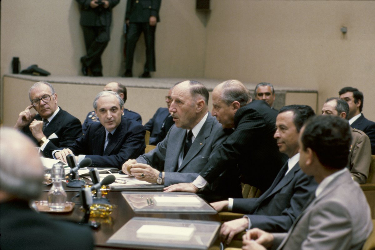 Several men in suits sitting in the NATO headquarters. One man is leaning over to another, seemingly saying something to him.