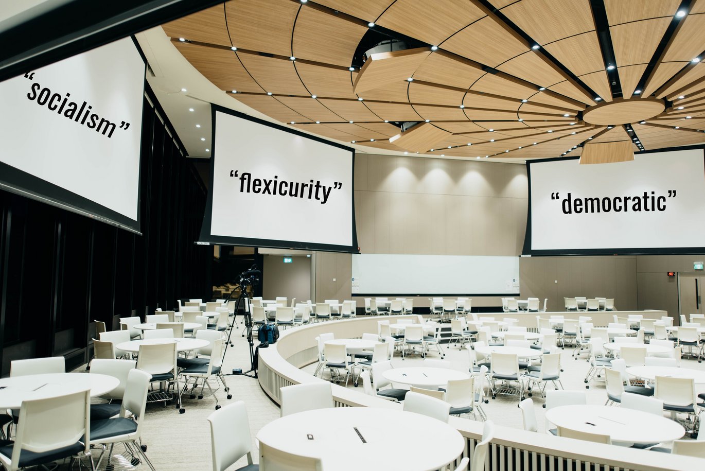 Large conference room with white upholstery with three large screens with "socialism", "flexicurity" and "democratic" on them.