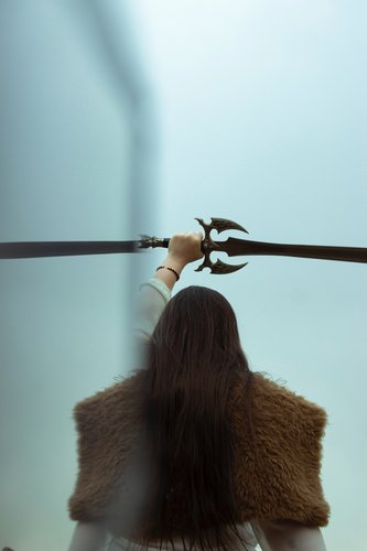 A picture of a woman seen from behind. She has long brown hair which is falling onto a big piece of fur worn as a coat. She is holding one hand above her head with a sword.