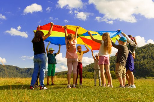 Pictured is a group of children who are standing in a circle on green grass, playing with a giant rainbow-coloured parachute between them. The sky is blue and everyone looks very happy. The photo is very colourful and lively.
