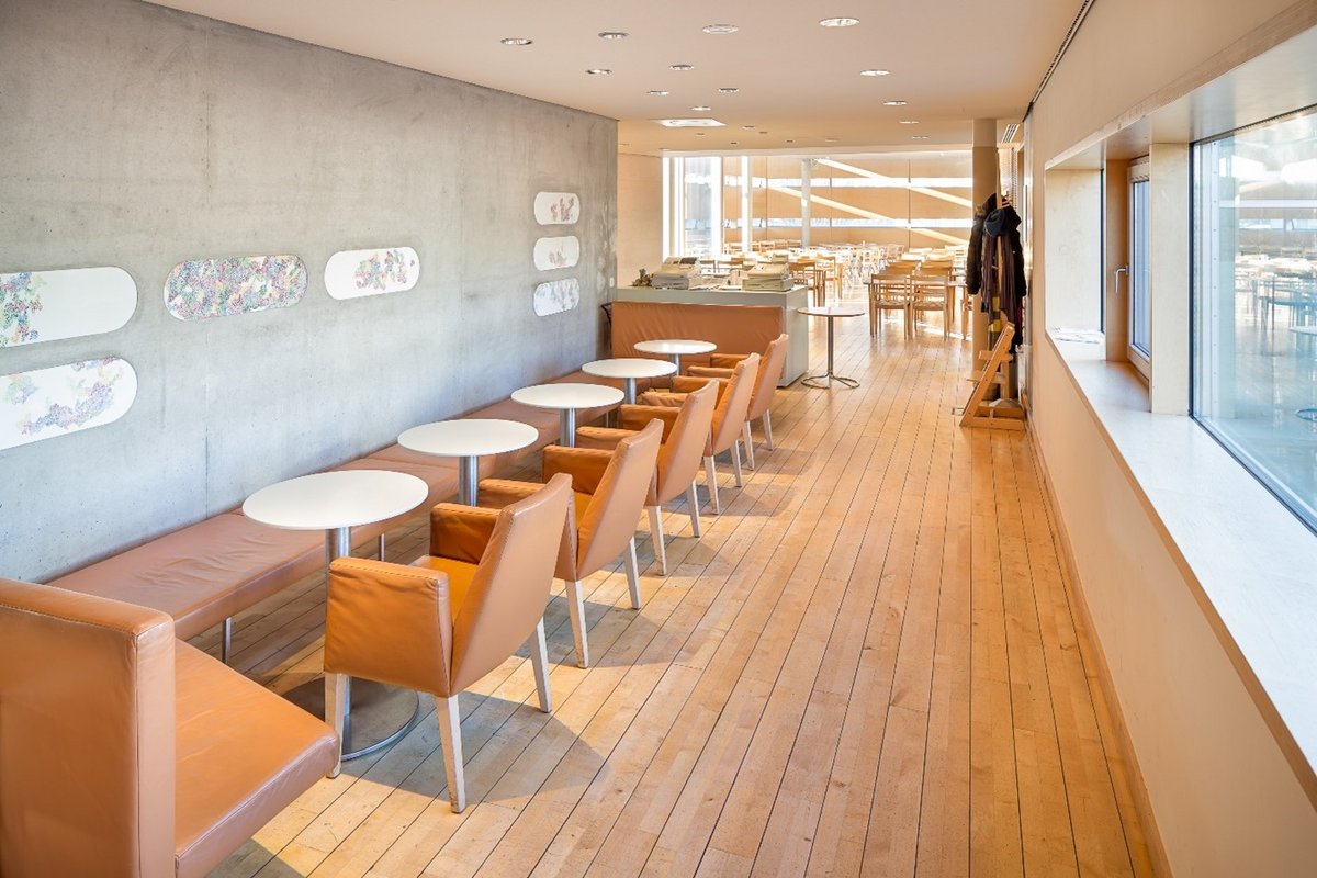 A photo of the inside of a building. There are bright wooden floors and modern sleek-looking tables and chairs along one wall. At the end of the room is a big wall of windows.