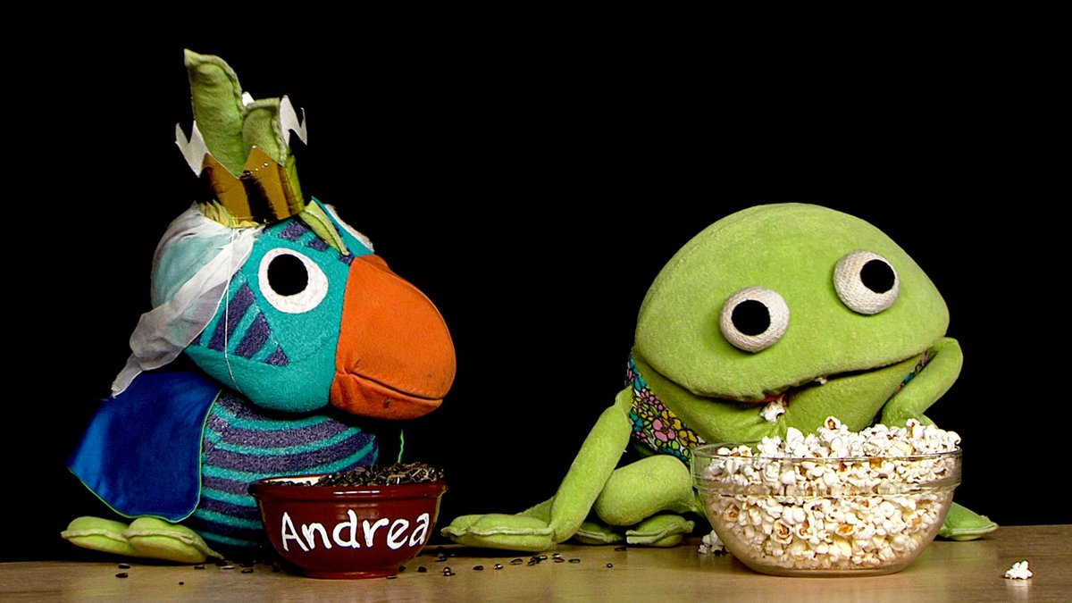 Pictured is two characters from the Danish children's show ‘Kaj and Andrea’. The two puppets are sitting besides each other. Andrea is a blue parrot wearing a princess hat and she is eating seeds. Kaj is a green frog and he is eating popcorn.