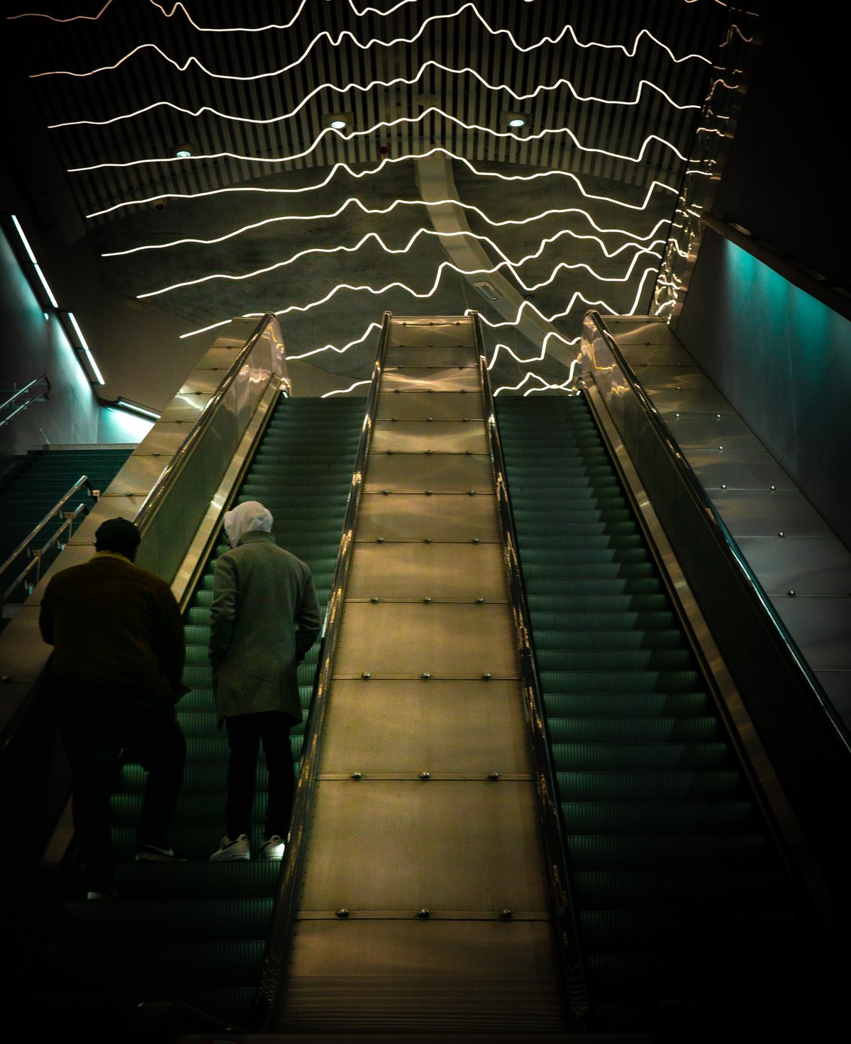An escalator in Stockholm seen from below. The ceiling is filled with lights in zig-zag shapes, a kind of art constellation. Two boys are standing on the escalator going upwards, wearing hats and hoodies with their back towards the camera. The photo is very dark.