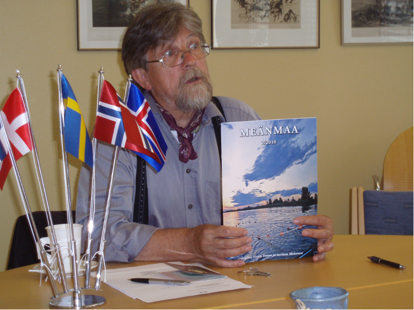 An elderly man 60+ year old sitting at a table holding a book with the title "MEÄNMAA". On the table is a collection of Nordic flags.