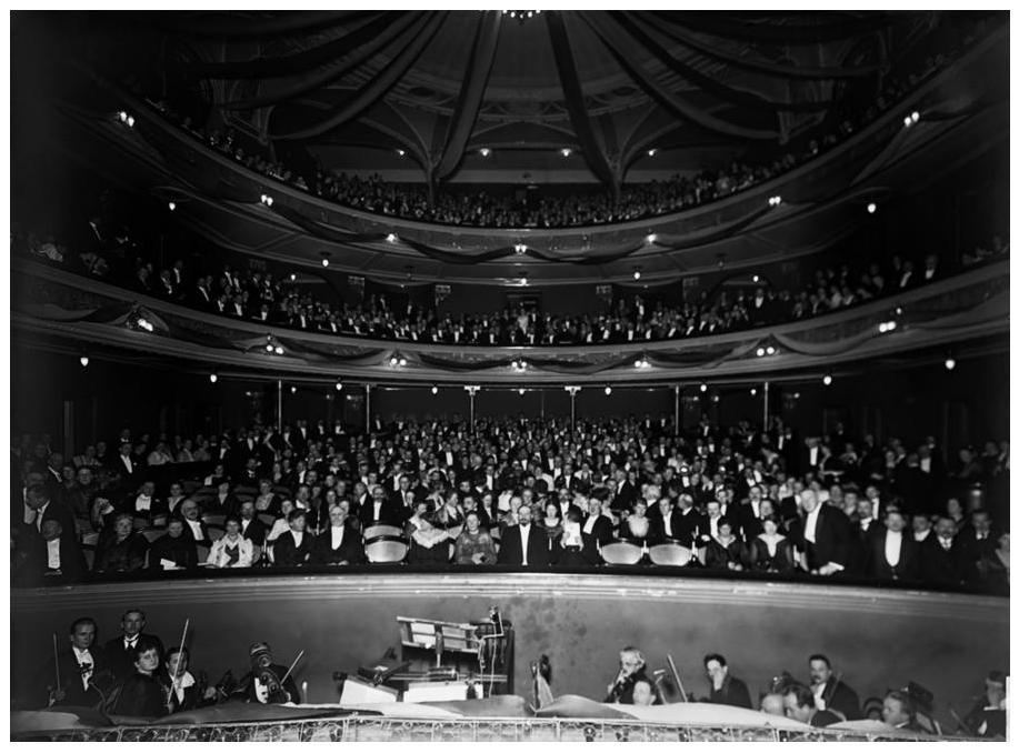 Inside the theatre with a large audience of people sitting, and looking towards the stage. At the bottom, you can spot the orchestra band playing
