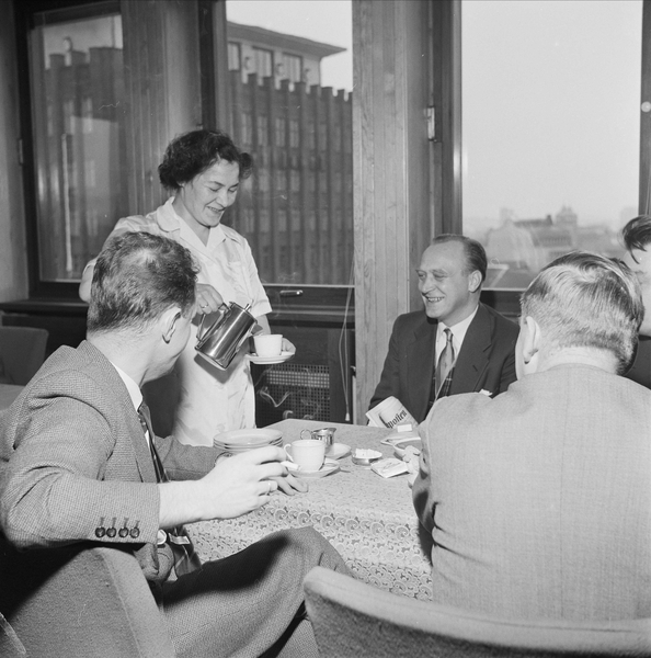 An old photograph of a woman serving a hot beverage of a group of men at a table.