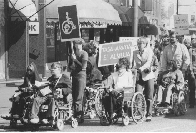 A big group of disabled pepple, some sitting in wheelchairs, others holding them or standing up, holding signs with the symbol of disability