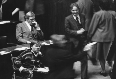 In a busy parliament, Kalle sits to the left alone, looks like he is intensely thinking.