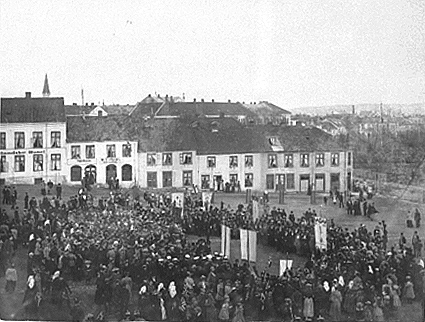 A public square filled with people, som carrying long tall flags, white buildings in the background