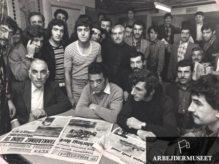 Black and white picture of a group of men standing and sitting with a newspaper on table in front