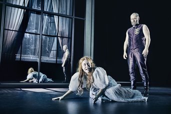 A dark room in which a woman is on the looking distraught while a man is standing behind her, looking down upon her. There is a mirror on the backwall, giving the stage an eerie feeling.