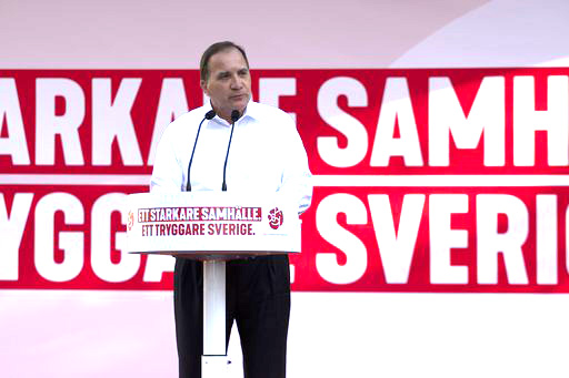 En bild på Sveriges statsminister Stefan Löfven. Han är klädd i svarta byxor och vit skjorta och står framför ett podium med två mikrofoner. Han ser allvarlig ut.