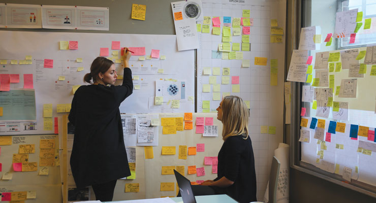Two female students communicating, one writing something on a board with many post-it notes and one sitting down.