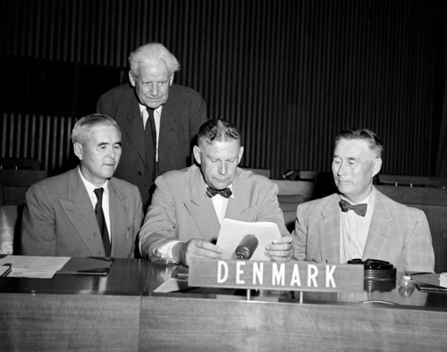 Three men sitting beside eachother at a table with the sign "DENMARK". Behind them stands an elderly man