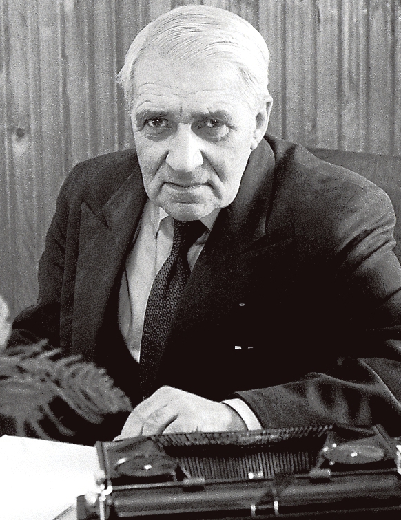 Man with white hair and a dark suit on next to a typewriter looking at the camera.
