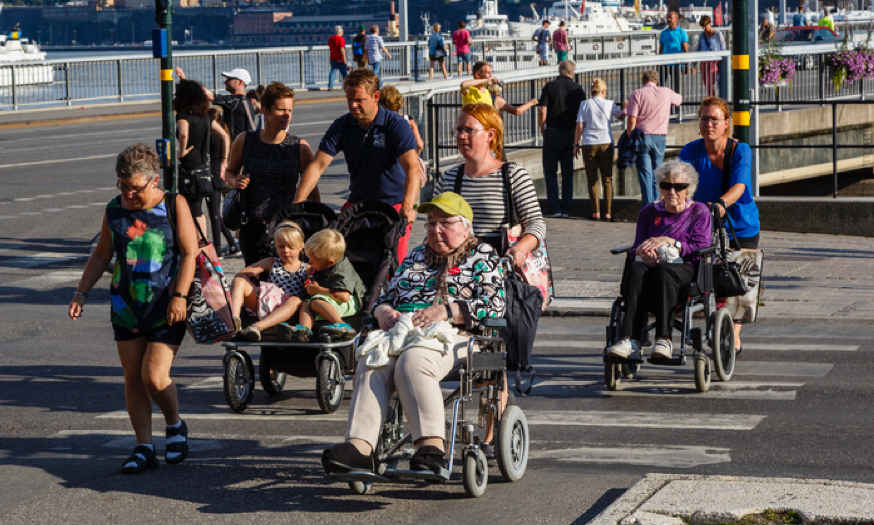 People of all ages walking on a bridge together. Two elderly people on wheelchairs.